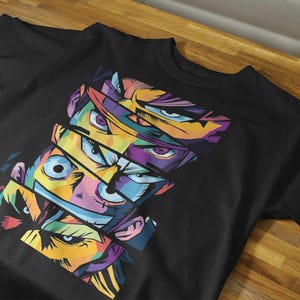 May include: Black t-shirt featuring a vibrant, pop art style design of multiple anime-style faces with colorful eyes and facial features. The design is composed of geometric shapes and bold colors, creating a striking visual effect.
