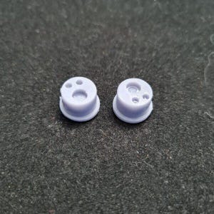 May include: Two small, light purple plastic parts with a circular base and a cylindrical top. Each part has a central hole and smaller holes around it. The parts are likely components of a larger product.