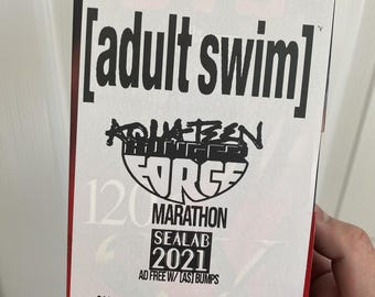 May include: A VHS tape with a red and black label. The label says "[adult swim]" and "SEALAB 2021" with the text "AD FREE W/ [AS] BUMPS" underneath. The tape is labeled "T-120 SX" and "6 HOURS".
