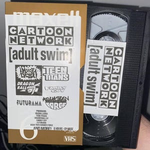 Puede incluir: Una cinta VHS con una etiqueta dorada que presenta el logotipo de Cartoon Network y el texto "adult swim". La etiqueta también incluye logotipos de varios programas, incluidos "Dragon Ball Z", "Teen Titans", "Family Guy", "Futurama" y "Aqua Teen Hunger Force". La cinta está etiquetada con el texto "246m" y "VHS".