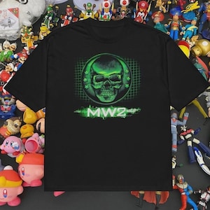Puede incluir: Camiseta negra con un diseño de calavera verde dentro de un círculo. El diseño incluye estrellas y el texto "MW2" en verde neón. La camiseta es de cuello redondo y manga corta.