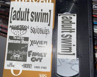 Könnte beinhalten: Ein goldfarbenes VHS-Band mit einem schwarzen Etikett. Das Etikett trägt den Text "[adult swim]" in großen weißen Buchstaben. Unter dem Text befinden sich mehrere kleinere Logos für Shows, darunter "Aqua Teen Hunger Force", "Squidbillies", "Tim and Eric Awesome Show", "Xavier: Renegade Angel", "Lucy, the Daughter of the Devil" und "Family Guy". Das Band trägt außerdem den Text "+MORE! 6 HOURS - EP MODE" und "VHS".