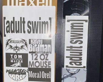 May include: A gold VHS tape with the text "maxell" and "[adult swim]" printed on the front. The tape features a collection of Adult Swim shows including "Metalocalypse", "Harvey Birdman, Attorney at Law", "Tom Goes to the Mayor", "12 oz Mouse", "Aqua Teen Hunger Force", and "Moral Orel". The tape also includes the text "+MOREJ 4 HOURS - EPMODE" and "VHS".