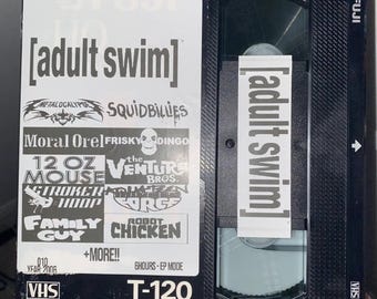 May include: A black VHS tape with a white label that says "adult swim" and features logos for various animated shows, including "Metalocalypse", "Squidbillies", "Moral Orel", "Frisky Dingo", "12 oz Mouse", "The Venture Bros.", "Stroker and Hoop", "The Brak Show", "Robot Chicken", and "Family Guy". The tape is labeled "T-120" and is 246m/807' long.