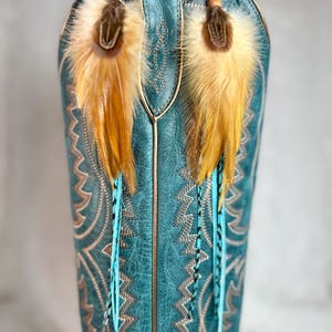 May include: A pair of feather earrings with a turquoise and brown color scheme. The earrings are hanging in front of a turquoise cowboy boot. The earrings have a brown top with yellow and brown feathers and turquoise and black feathers.