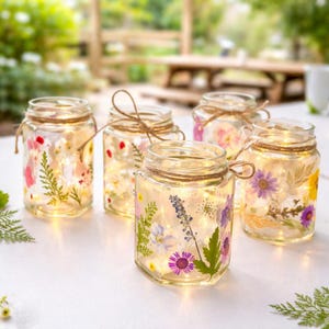 May include: Five glass jars filled with dried flowers and fairy lights. Each jar has twine tied around the rim. The flowers are in shades of pink, purple, and yellow. The jars are arranged on a white surface.