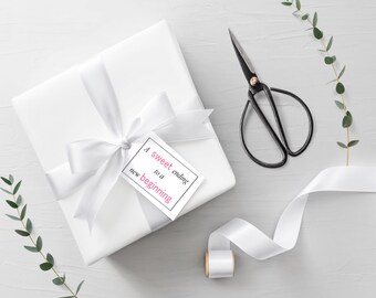 Può includere: Una confezione regalo bianca legata con un nastro di raso, un'etichetta con il testo "A sweet ending to a new beginning", forbici nere e un rotolo di nastro bianco sono disposti su una superficie grigio chiaro. Dei rametti verdi aggiungono un tocco naturale.