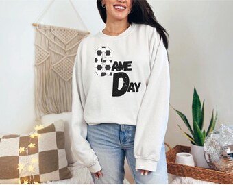 Puede incluir: Una sudadera blanca con las palabras "GAME DAY" en letras negras y gráficos de balones de fútbol. La sudadera es de cuello redondo y se usa con vaqueros azules. El tema general es el deporte.