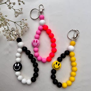 Könnte beinhalten: Drei Perlen-Schlüsselanhänger mit Smiley-Gesichtern. Der Schlüsselanhänger links hat weiße und schwarze Perlen mit einem schwarzen Smiley-Gesicht. Der Schlüsselanhänger in der Mitte hat rosa und rote Perlen mit einem rosa Smiley-Gesicht. Der Schlüsselanhänger rechts hat gelbe und schwarze Perlen mit einem gelben Smiley-Gesicht.