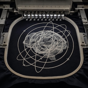 May include: A black fabric piece secured in an embroidery hoop featuring a white stitched design of a human brain surrounded by intersecting orbital rings with small star-like accents. The embroidery machine needles are positioned above.