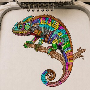 May include: An embroidery design of a chameleon perched on a brown branch, featuring a vibrant, multi-colored geometric pattern with shades of blue, green, yellow, orange, and pink, secured within a metal embroidery hoop on white fabric.