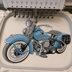 Puede incluir: Un diseño de bordado de una motocicleta azul claro con neumáticos de banda blanca, asiento negro y alforjas negras cosido sobre una tela color crema sujeta en un bastidor de bordado rectangular bajo una máquina de bordar de varias agujas.