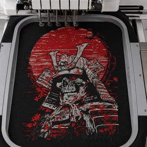 May include: An embroidery machine hoop holds black fabric featuring a detailed embroidered design of a skeletal samurai warrior wearing traditional armor. The artwork includes a large red circular sun background and small red floral accents.