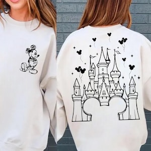May include: White sweatshirt featuring a black and white Mickey Mouse graphic on the front and a castle design on the back. The sweatshirt has a crew neck and long sleeves. The design includes Mickey Mouse, a castle, and balloons.