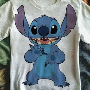 Può includere: T-shirt bianco girocollo con un personaggio dei cartoni animati blu. Il personaggio ha grandi orecchie, un ampio sorriso e occhi scuri. È in posa giocosa, con le mani vicino al viso. La maglietta è realizzata in materiale morbido.
