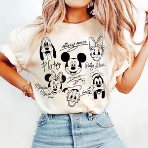 May include: A white t-shirt with black and white illustrations of Mickey Mouse, Minnie Mouse, Donald Duck, Goofy, Pluto, and Daisy Duck. The characters are drawn in a cartoon style and have their names written next to them.