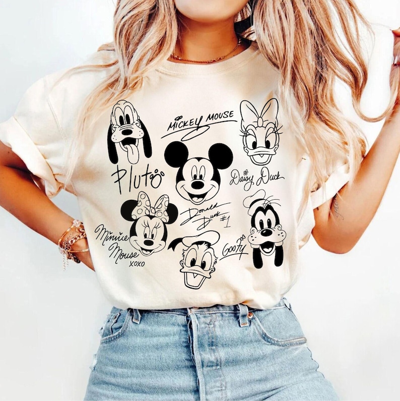 May include: A white t-shirt with black and white illustrations of Mickey Mouse, Minnie Mouse, Donald Duck, Goofy, Pluto, and Daisy Duck. The characters are drawn in a cartoon style and have their names written next to them.