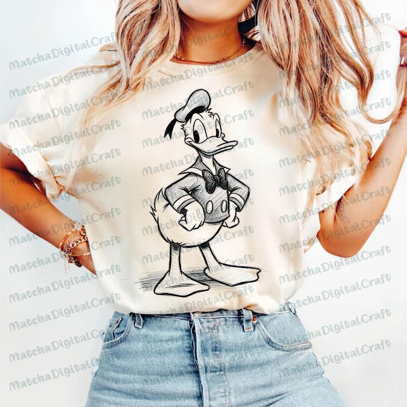 K&ouml;nnte beinhalten: Cremefarbenes T-Shirt mit einer Schwarz-Wei&szlig;-Illustration von Donald Duck in Matrosenkleidung. Die Zeichentrickfigur ist mit verschr&auml;nkten Armen dargestellt und tr&auml;gt eine Matrosenm&uuml;tze und eine Fliege. Das Shirt ist ein Freizeitartikel.
