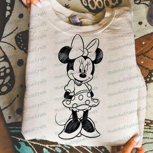 K&ouml;nnte beinhalten: Ein hellbeige Sweatshirt mit einer schwarz-wei&szlig;en Skizze von Minnie Mouse. Die Cartoonfigur tr&auml;gt ein gepunktetes Kleid und eine gro&szlig;e Schleife. Das Sweatshirt ist gefaltet und wird von H&auml;nden gehalten.