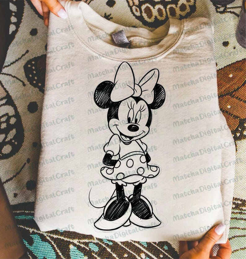 K&ouml;nnte beinhalten: Ein hellbeige Sweatshirt mit einer schwarz-wei&szlig;en Skizze von Minnie Mouse. Die Cartoonfigur tr&auml;gt ein gepunktetes Kleid und eine gro&szlig;e Schleife. Das Sweatshirt ist gefaltet und wird von H&auml;nden gehalten.
