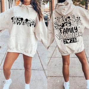 May include: Cream-colored hoodie featuring a black and white graphic of Disney characters on the front and back. The front displays Mickey, Minnie, and friends. The back has the text "FAMILY VACATION 2026" with a Mickey Mouse head.