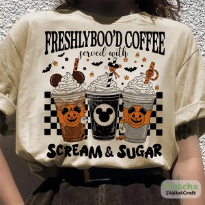 May include: Beige t-shirt with a Halloween design. It shows three coffee cups with whipped cream, pumpkin faces, and the text "FRESHLYBOO'D COFFEE served with SCREAM & SUGAR". Bats, stars, and candy corn decorate the design.