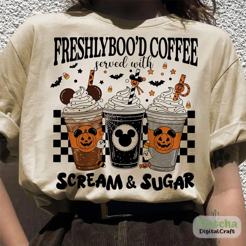 May include: Beige t-shirt with a Halloween design. It shows three coffee cups with whipped cream, pumpkin faces, and the text "FRESHLYBOO'D COFFEE served with SCREAM & SUGAR". Bats, stars, and candy corn decorate the design.