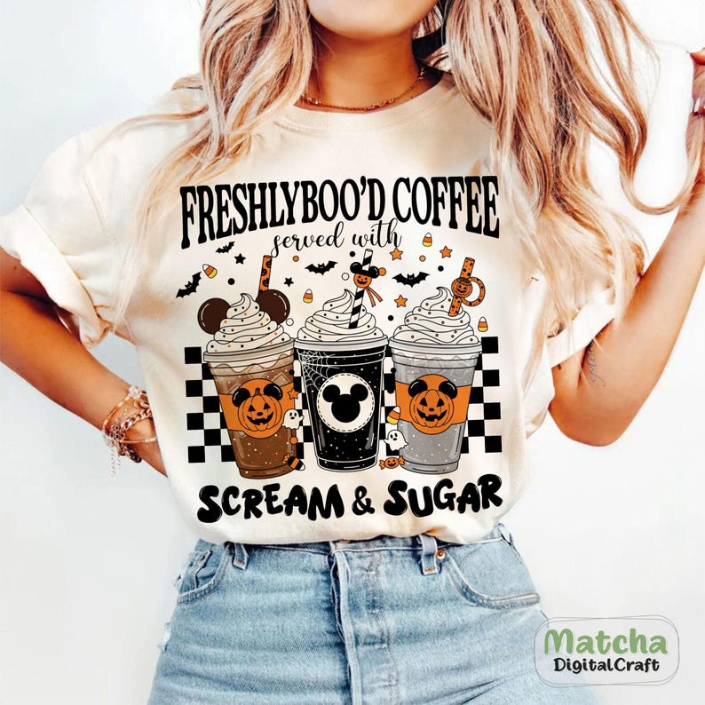 May include: A cream-coloured t-shirt with a Halloween design. The graphic shows three coffee cups with whipped cream, pumpkin and Mickey Mouse designs, and the text "FRESHLYBOO'D COFFEE served with SCREAM & SUGAR."
