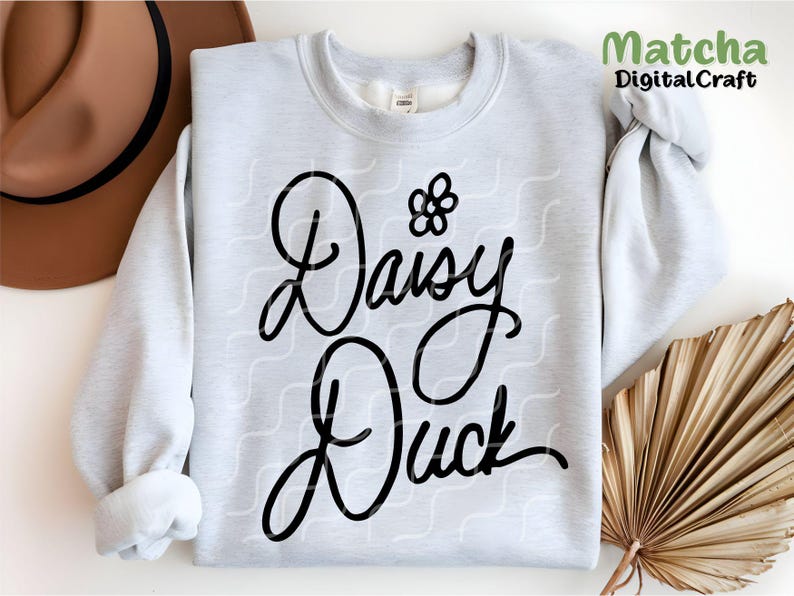 Puede incluir: Una sudadera blanca con un dise&ntilde;o gr&aacute;fico negro que dice "Daisy Duck" con una peque&ntilde;a flor sobre la palabra "Daisy".