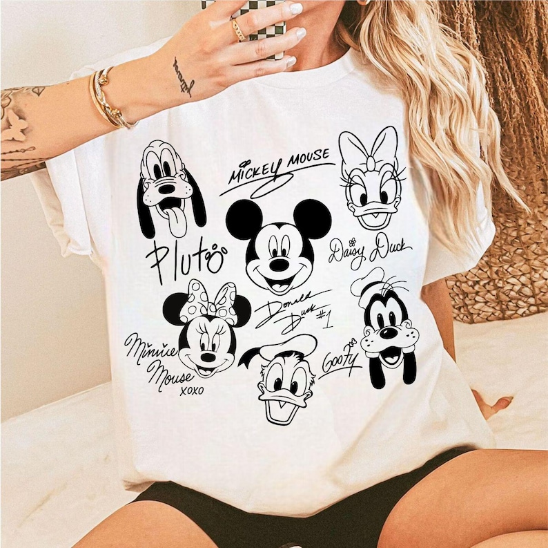 May include: White t-shirt with black and white cartoon images of Mickey Mouse, Minnie Mouse, Pluto, Goofy, Donald Duck, and Daisy Duck. Each character has their name written in cursive next to them.