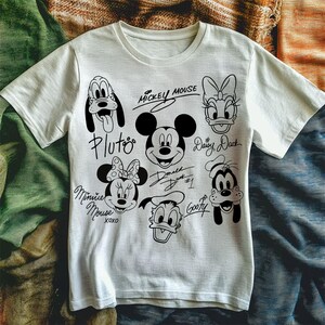 May include: White t-shirt with black and white illustrations of Mickey Mouse, Minnie Mouse, Pluto, Goofy, Donald Duck, and Daisy Duck. Each character has their signature underneath. The text reads "Mickey Mouse", "Pluto", "Minnie Mouse XOXO", "Donald Duck #1", "Goofy", and "Daisy Duck".