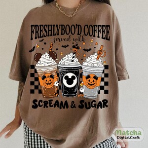 May include: Brown t-shirt with a Halloween design. The graphic shows three coffee cups with whipped cream, jack-o'-lanterns, and the text "FRESHLYBOO'D COFFEE served with SCREAM & SUGAR". Bats and candy corn are also included in the design.