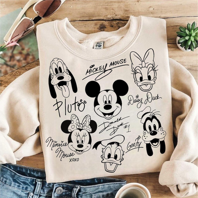 May include: A beige sweatshirt with black and white illustrations of Mickey Mouse, Minnie Mouse, Donald Duck, Daisy Duck, Goofy, and Pluto. The characters are drawn in a cartoon style and have their names written next to them.