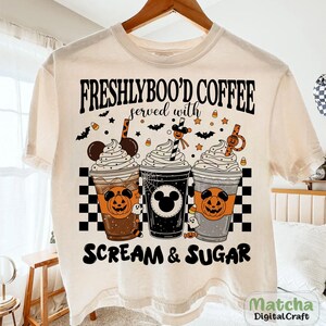 May include: Cream-coloured t-shirt with a Halloween design. The shirt features the text "FRESHLYBOO'D COFFEE served with SCREAM & SUGAR" above three illustrated coffee cups with whipped cream, Halloween decorations, and Mickey Mouse silhouettes.