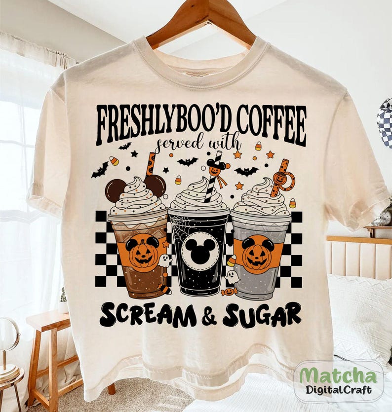 May include: Cream-coloured t-shirt with a Halloween design. The shirt features the text "FRESHLYBOO'D COFFEE served with SCREAM & SUGAR" above three illustrated coffee cups with whipped cream, Halloween decorations, and Mickey Mouse silhouettes.