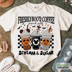 May include: Cream-coloured t-shirt with the text "FRESHLYBOO'D COFFEE served with SCREAM & SUGAR." The graphic shows three Halloween-themed coffee cups with whipped cream, bats, and sweets. The cups have pumpkin and Mickey Mouse designs.
