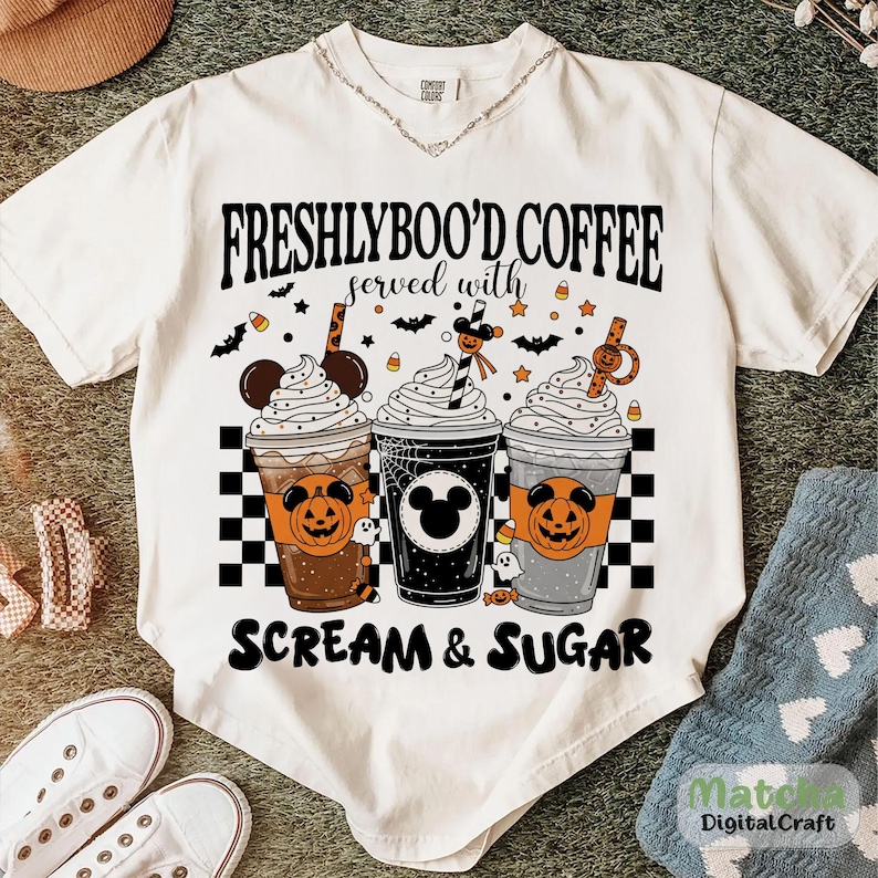 May include: Cream-coloured t-shirt with the text "FRESHLYBOO'D COFFEE served with SCREAM & SUGAR." The graphic shows three Halloween-themed coffee cups with whipped cream, bats, and sweets. The cups have pumpkin and Mickey Mouse designs.