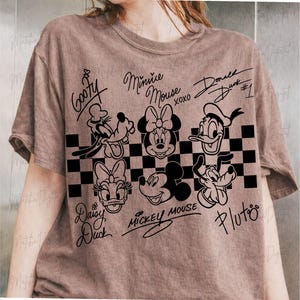 May include: A brown t-shirt featuring black line art of Disney characters including Goofy, Minnie Mouse, Donald Duck, Daisy Duck, Mickey Mouse, and Pluto. The characters are arranged above a black and white checkered pattern.