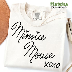 Puede incluir: Una camiseta blanca con un gr&aacute;fico negro que dice "Minnie Mouse xoxo" en una fuente cursiva. Hay dos peque&ntilde;os corazones sobre la palabra "Minnie".
