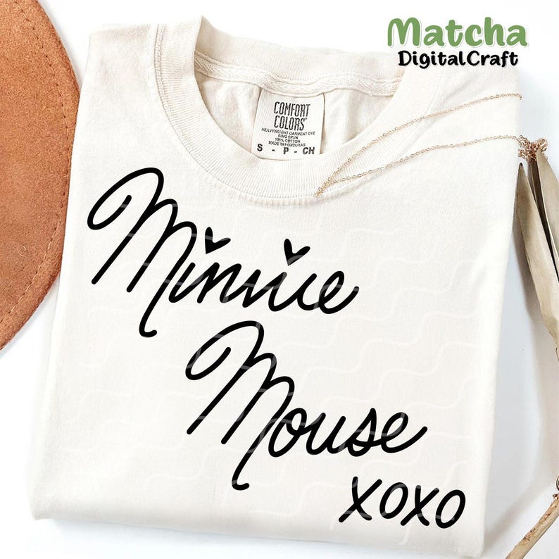 Puede incluir: Una camiseta blanca con un gr&aacute;fico negro que dice "Minnie Mouse xoxo" en una fuente cursiva. Hay dos peque&ntilde;os corazones sobre la palabra "Minnie".