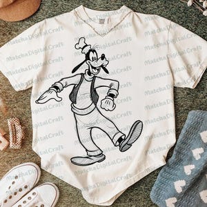 K&ouml;nnte beinhalten: Cremefarbenes T-Shirt mit einer Schwarz-Wei&szlig;-Skizze von Goofy in Bewegung. Das Shirt hat einen Rundhalsausschnitt und kurze &Auml;rmel. Eine silberne Halskette ist sichtbar. Der Text "Matcha Digital Craft" ist auf dem Shirt aufgedruckt.
