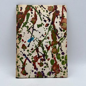 May include: A rectangular book with a cream-colored cover, adorned with a vibrant abstract design. Splatters and streaks of red, green, purple, and blue paint create a dynamic and colorful pattern across the surface. The book's edges are visible.