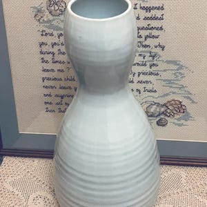 May include: A pale blue ceramic vase with a wide base and a narrow neck. The vase features a textured, ribbed design and a smooth, rounded top. The background includes a framed cross-stitch with text.