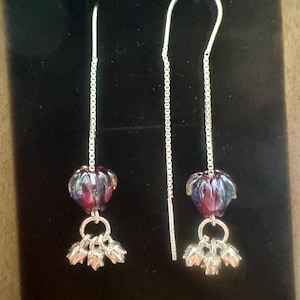 May include: A pair of dangle earrings with a silver chain and hook. Each earring features a purple and red glass bead and a cluster of silver flower charms. The earrings are displayed against a black background.