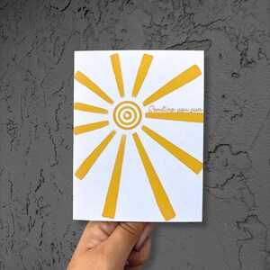 May include: A white greeting card featuring a yellow sunburst design. The sun has a circular centre with radiating lines. The words "Sending you sun" are written across the sunburst. The card is held by a hand against a textured grey background.