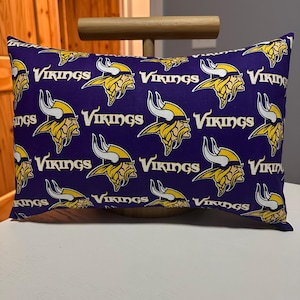 May include: Rectangular pillow with a purple background and a repeating pattern of the word "Vikings" and a yellow and white Viking head logo. The pillow is resting on a white surface.