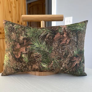 May include: Rectangular decorative pillow with a woodland camouflage pattern. The pillow features a detailed print of pine needles, pine cones, and fallen leaves in shades of green, brown, and tan. The pillow is displayed on a wooden stand.
