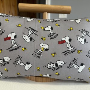 May include: Gray rectangular pillow featuring Snoopy from Peanuts in various poses with words like "Hungry", "Happy", and "Love". The pillow has a light gray background with yellow accents.