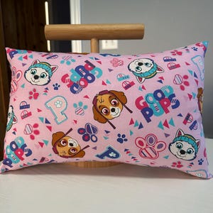 May include: Pink rectangular pillow featuring cartoon dog characters, paw prints, and the word "GOOD" in blue and red. The pillow has a repeating pattern of the characters and other design elements.
