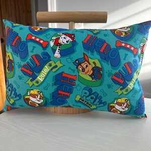 May include: Rectangular turquoise pillow featuring a repeating pattern of cartoon characters, the words "HERO PUPS", and paw prints. The pillow has a red border and is resting on a white surface.